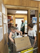 A young boy leans over a metal shopping cart filled with cardboard boxes. A man in a green shirt points toward the shelves of a food pantry, guiding the students as they organize supplies.